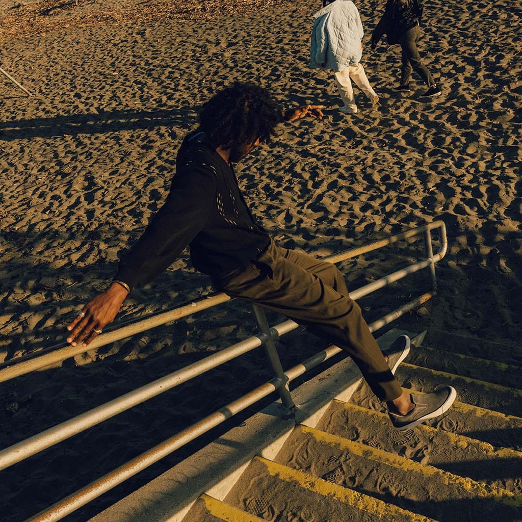 Men's Hawthorne Slip On in Pavement suede, worn by a person leaping on beach stairs at dusk