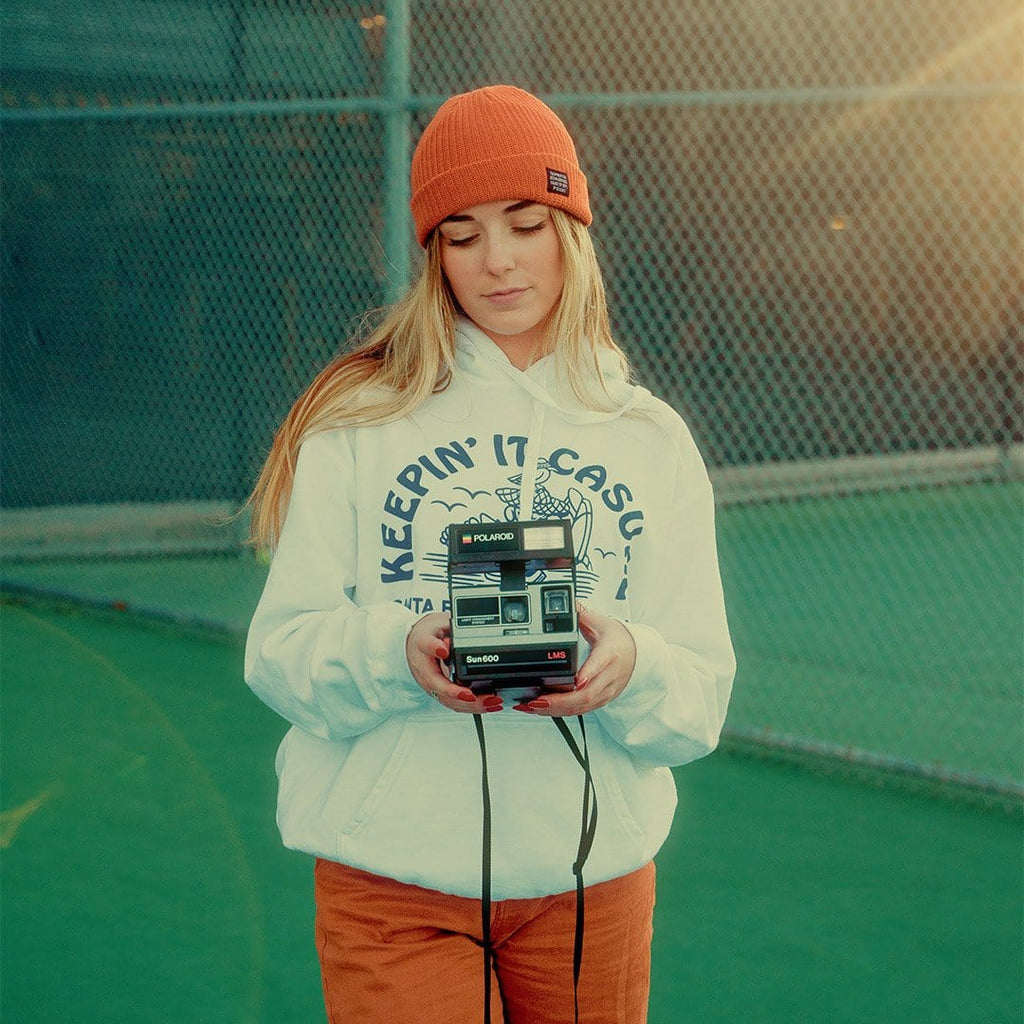 Person in a white SeaVees Keepin' It Casual hoodie and orange beanie holding a camera