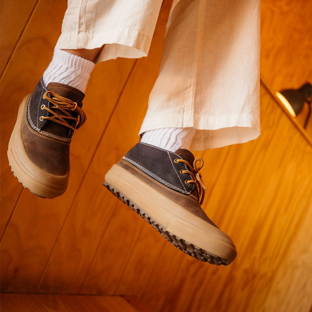 Women's Carson 3 Eye Boot in Peacoat Navy/Bark leather, worn with cream pants and white socks, feet raised, showing the lug