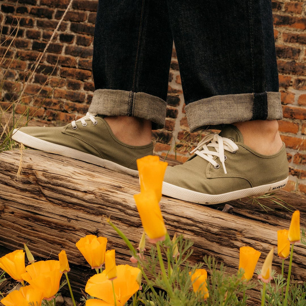 Men's Sixty Six Sneaker in Surplus Green canvas, close-up worn with cuffed jeans on a log beside orange flowers