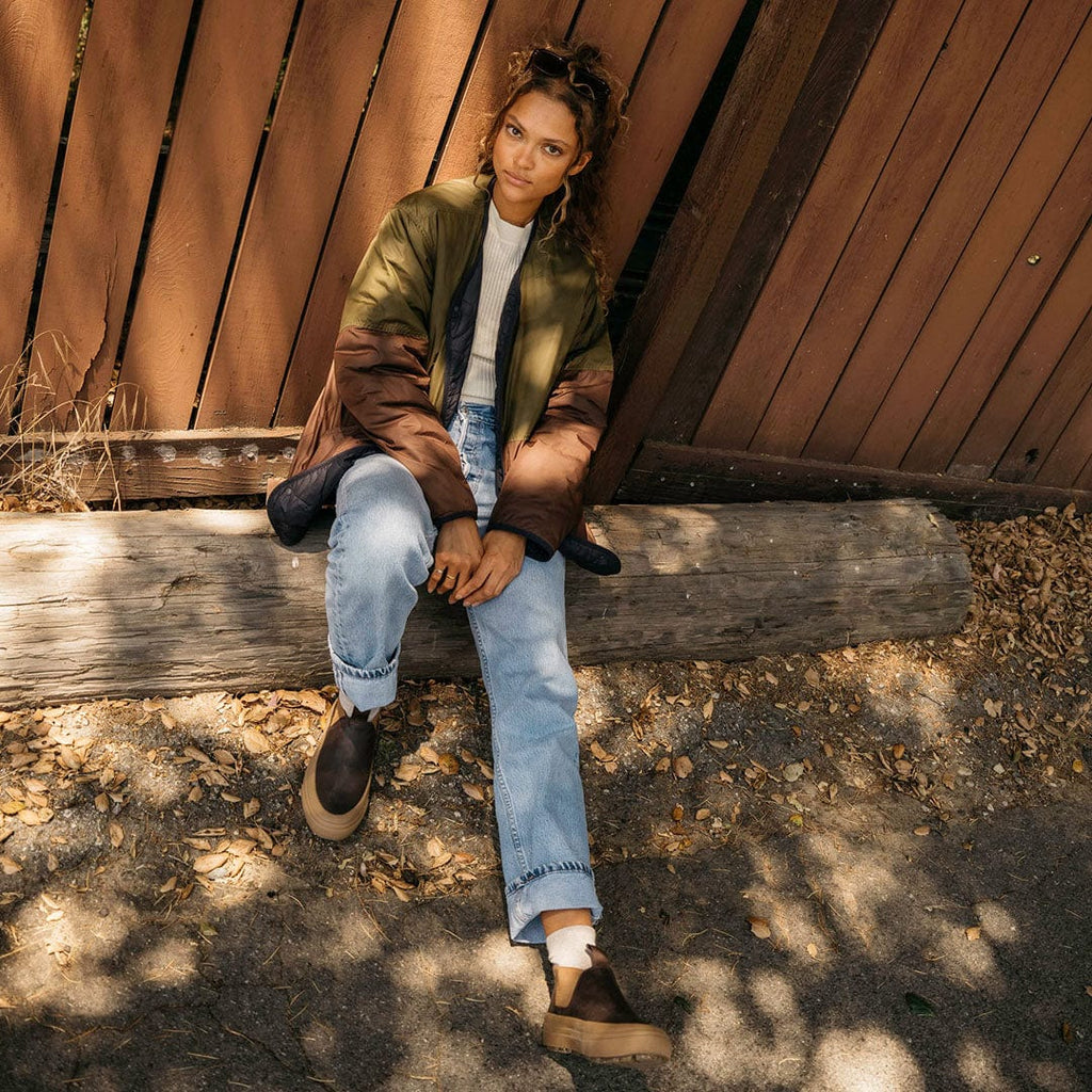 Women's Ballard Boot in Coffee Bean leather, worn by a person seated on a wooden bench outdoors
