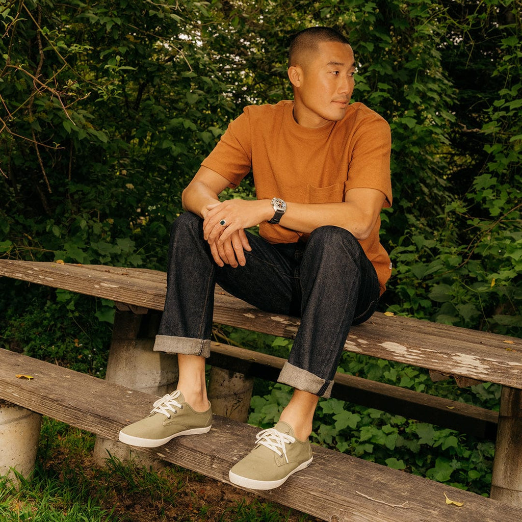 Men's Sixty Six Sneaker in Surplus Green canvas, worn with dark jeans by a person seated on a wooden bench