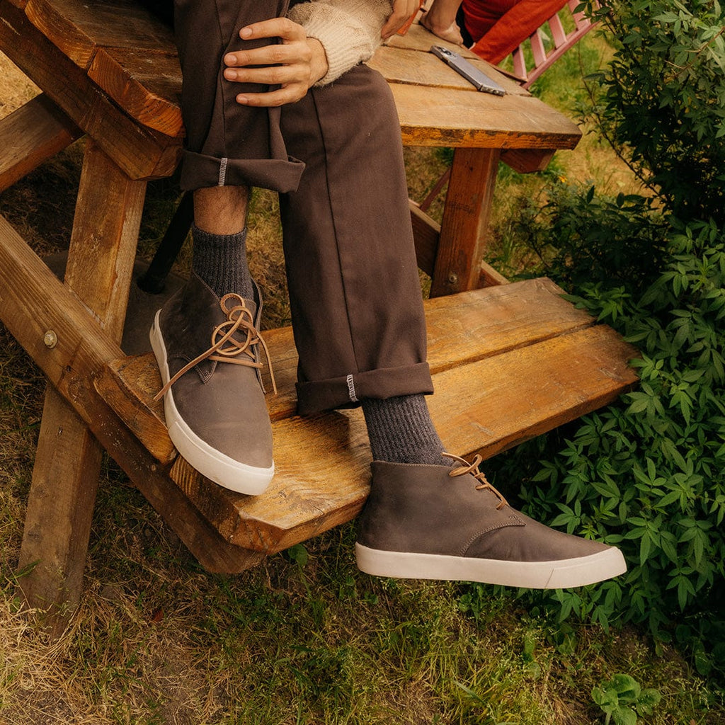 Men's Maslon Desert Boot in Charcoal leather, worn with brown pants by a person seated at a wooden picnic table