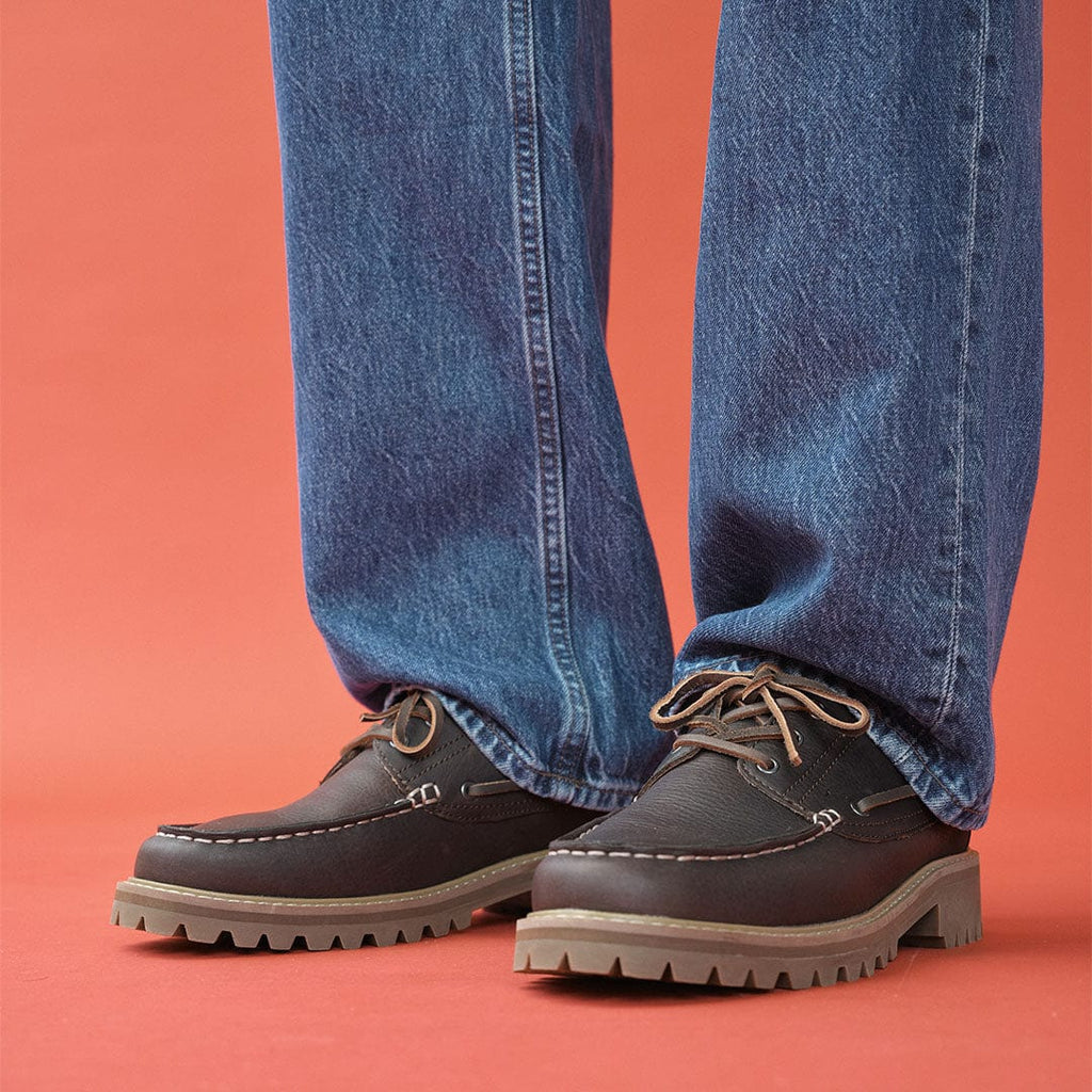 Men's Larkspur Lug Boat Shoe in Dark Chocolate leather, worn with wide-leg jeans on a coral background, close up