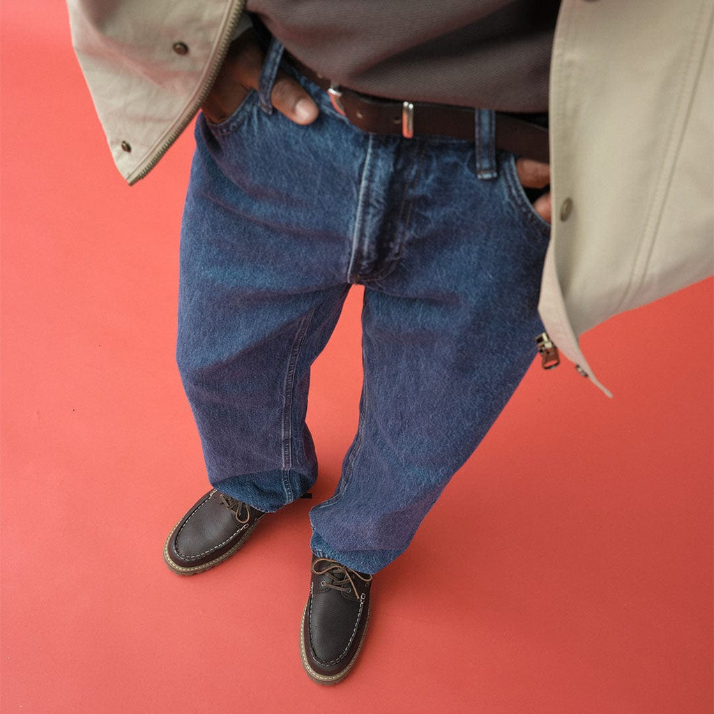 Men's Larkspur Lug Boat Shoe in Dark Chocolate leather, worn with jeans, standing on a coral background