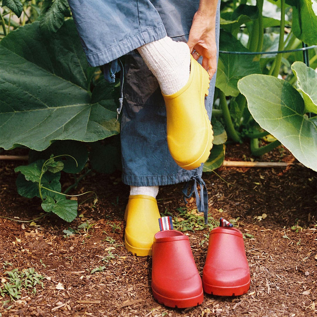 Women's Bodega Clog in Calendula rubber, worn with white socks and jeans in a garden, beside a red pair