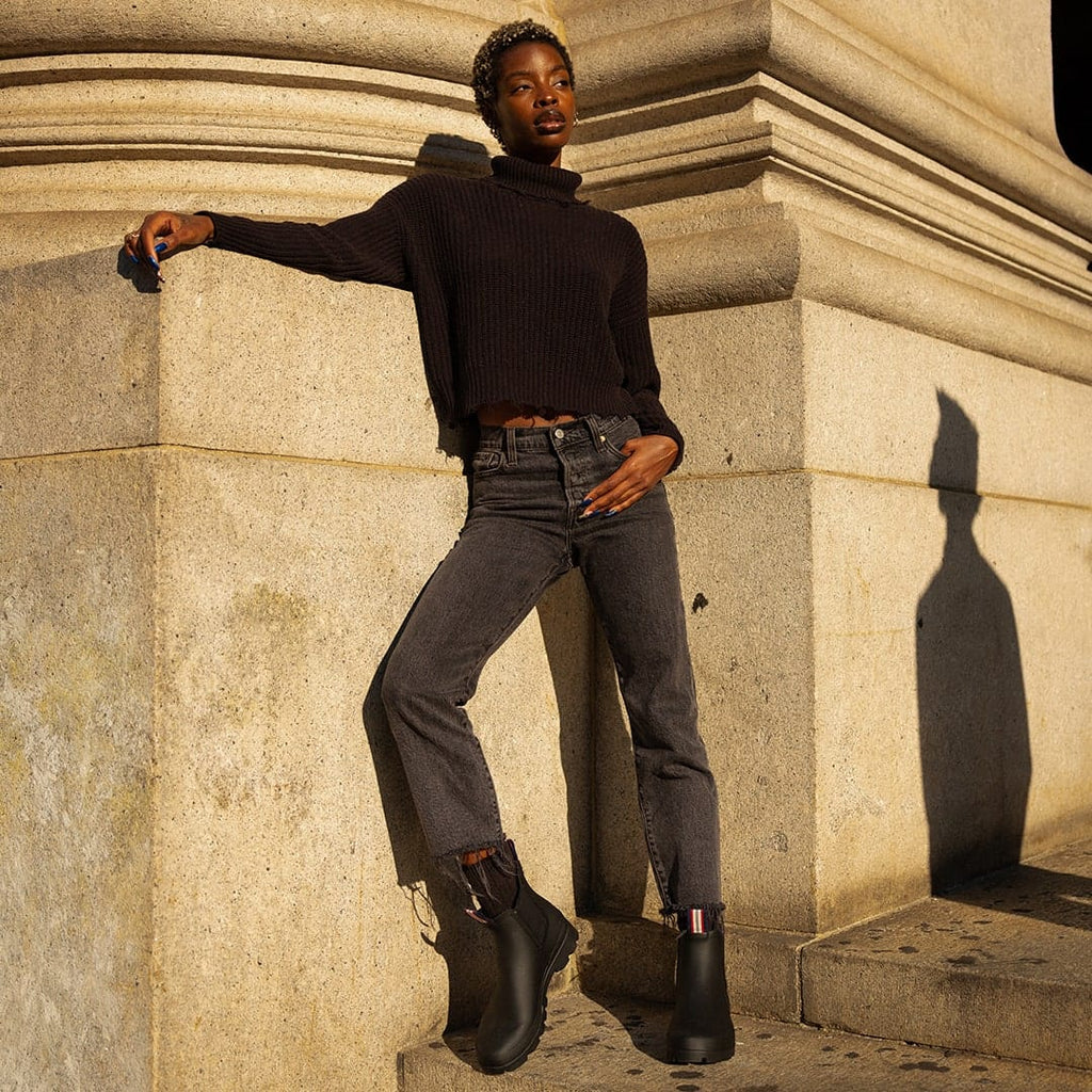Women's Bolinas Off Shore Boot in Black rubber, worn with dark pants by a person leaning on a stone column