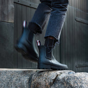 Women's Bolinas Off Shore Boot in Black rubber, close-up worn with dark jeans stepping on a rock
