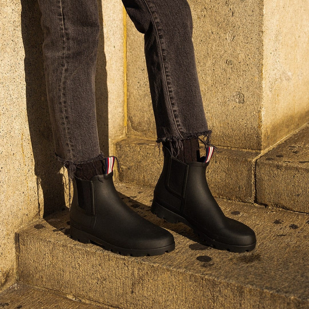 Women's Bolinas Off Shore Boot in Black rubber, close-up worn with dark jeans on stone steps