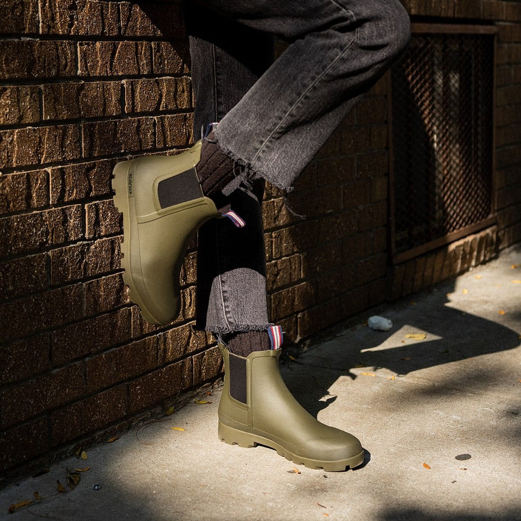 Women's Bolinas Off Shore Boot in Military Olive rubber, close-up worn with gray pants against a brick wall