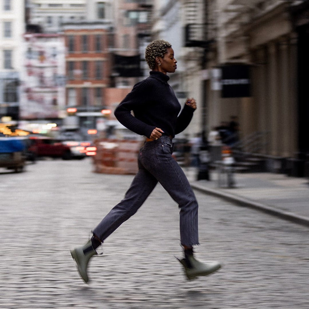 Women's Bolinas Off Shore Boot in Military Olive rubber, worn by a person running down a cobblestone city street