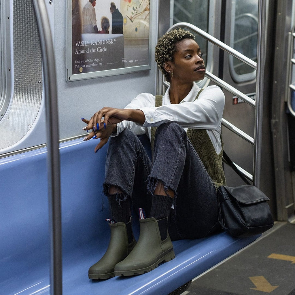 Women's Bolinas Off Shore Boot in Military Olive rubber, worn with dark pants by a person seated on a subway bench