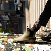 Women's Bolinas Off Shore Boot in Military Olive rubber, close-up worn with dark pants on a city sidewalk