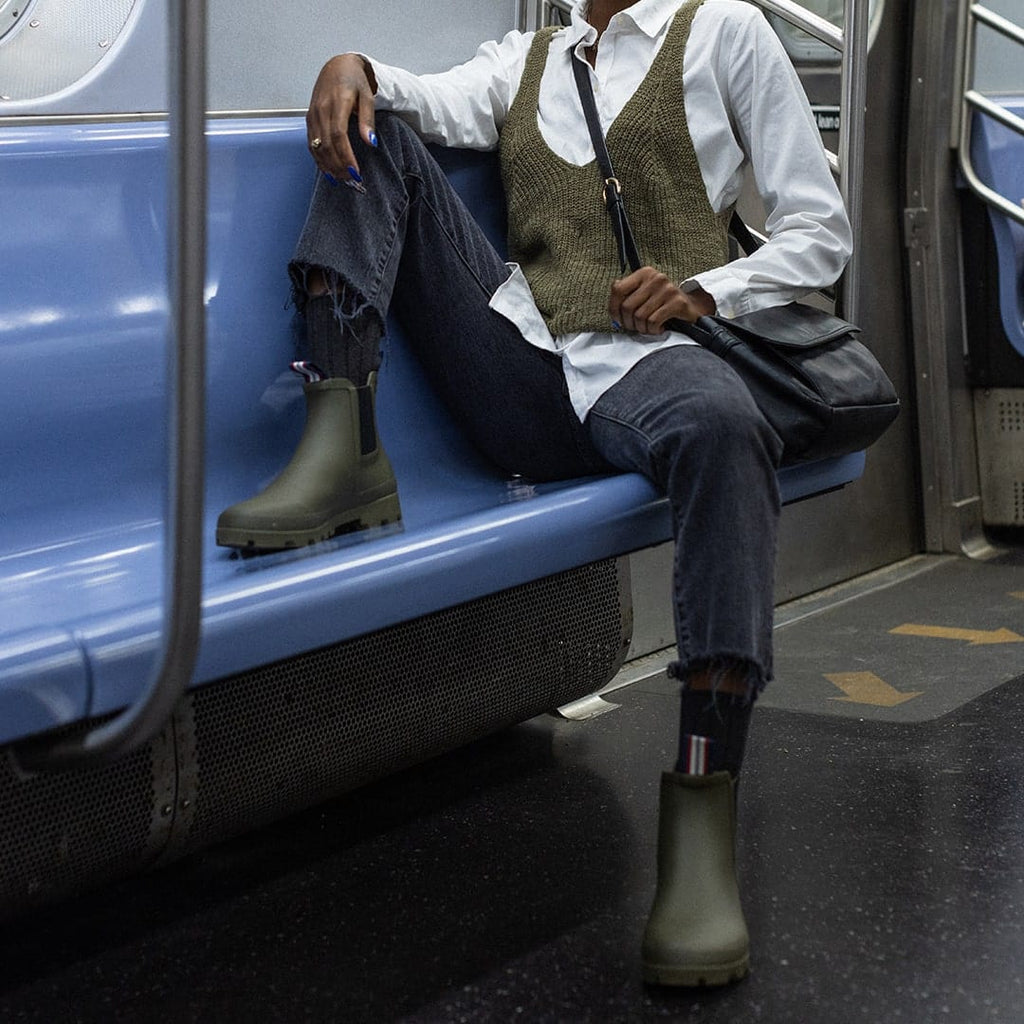 Women's Bolinas Off Shore Boot in Military Olive rubber, worn by a person seated on a subway bench