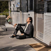 Men's Hermosa Sneaker in Black canvas, worn with a dark suit by a person seated against a trailer wall