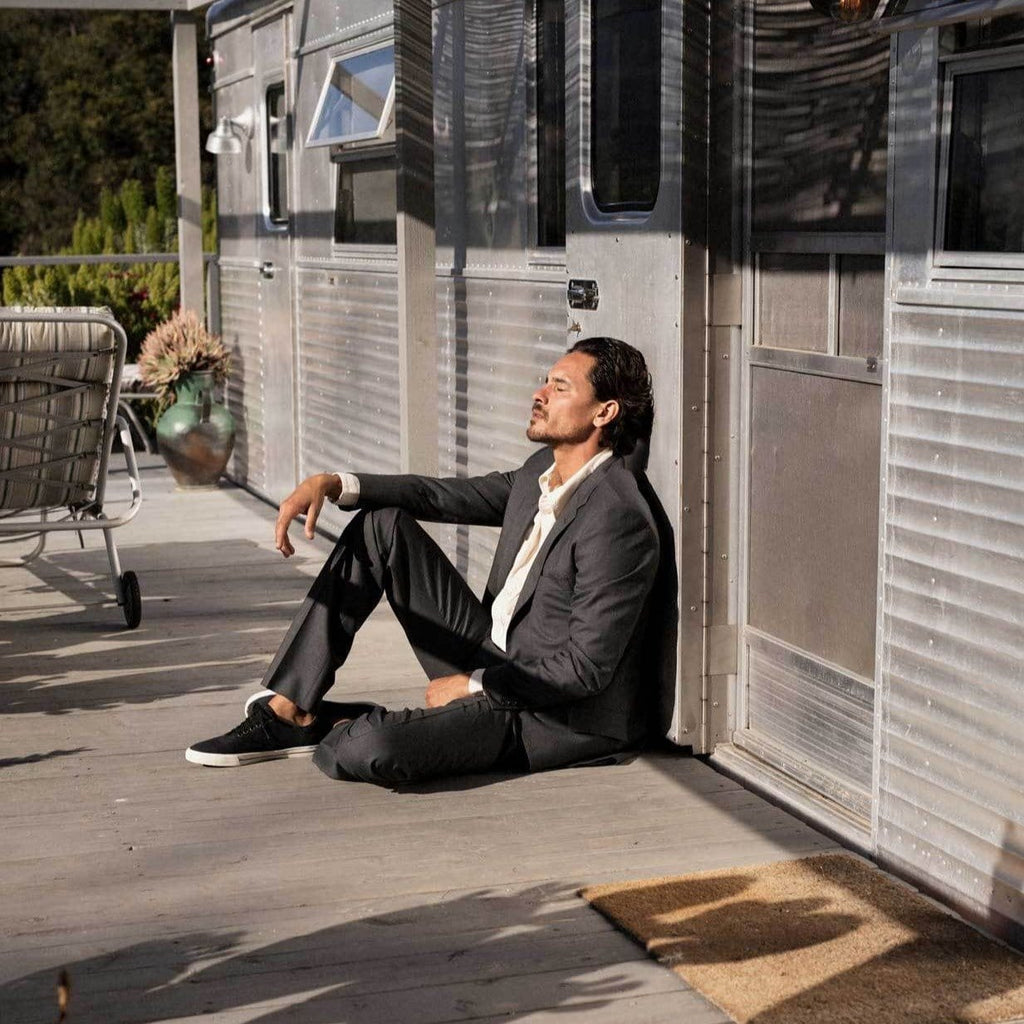 Men's Hermosa Sneaker in Black canvas, worn with a dark suit by a person seated against a trailer wall