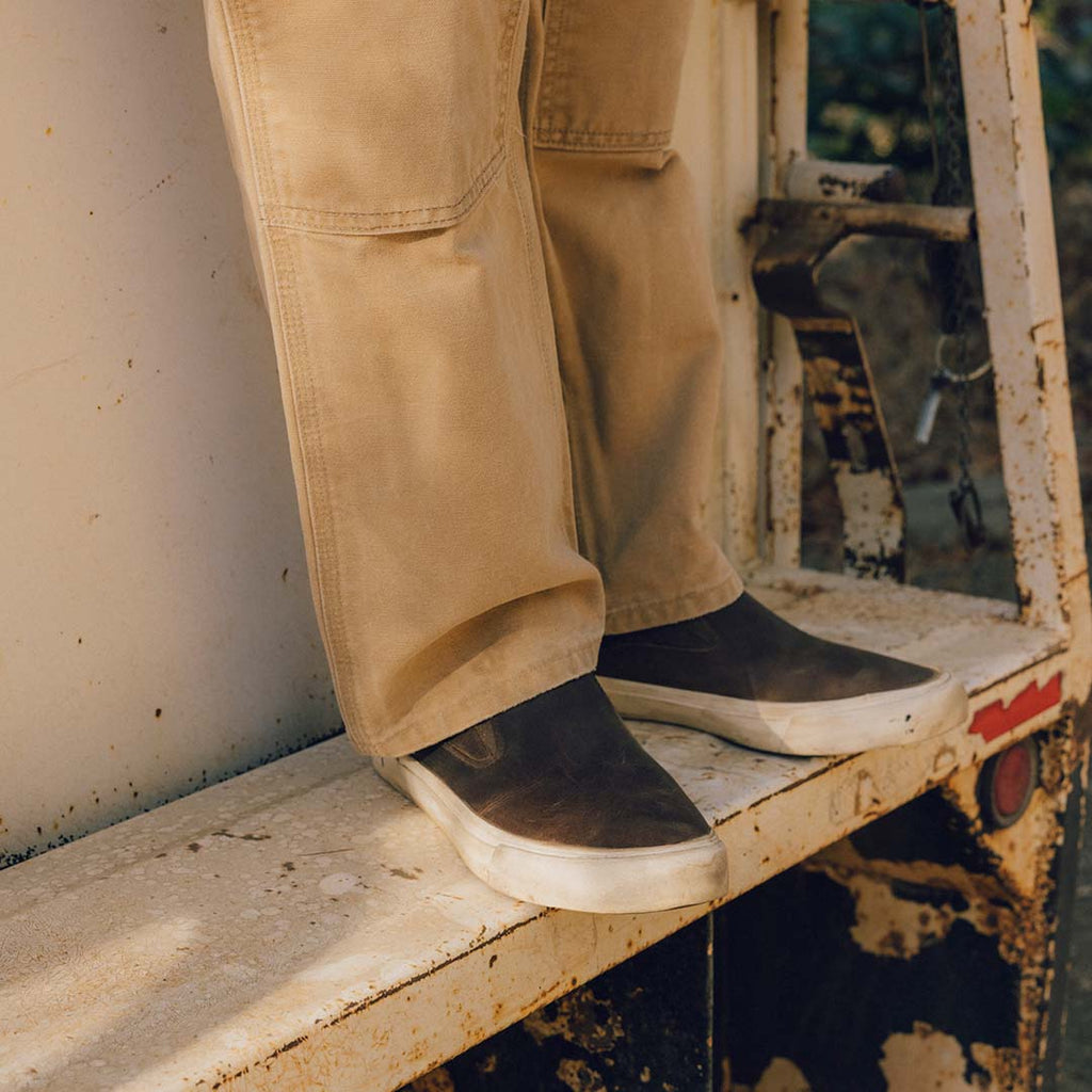 Men's Hawthorne Slip On in Elmwood leather, worn with khaki pants on a wooden ledge