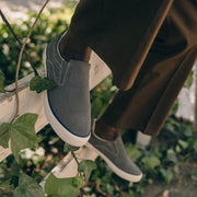 Men's Hawthorne Slip On in Pavement suede, close-up worn with brown pants among green leaves