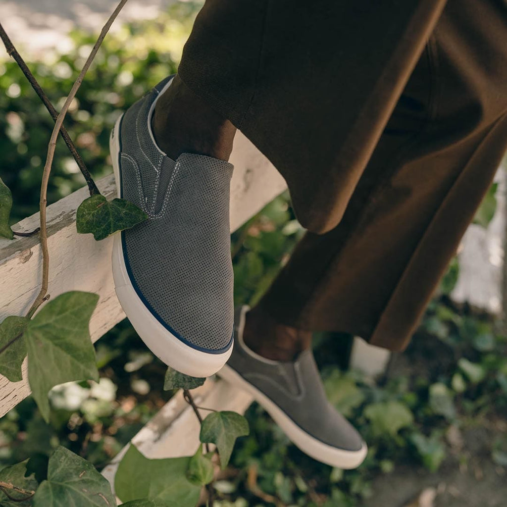 Men's Hawthorne Slip On in Pavement suede, close-up worn with brown pants among green leaves
