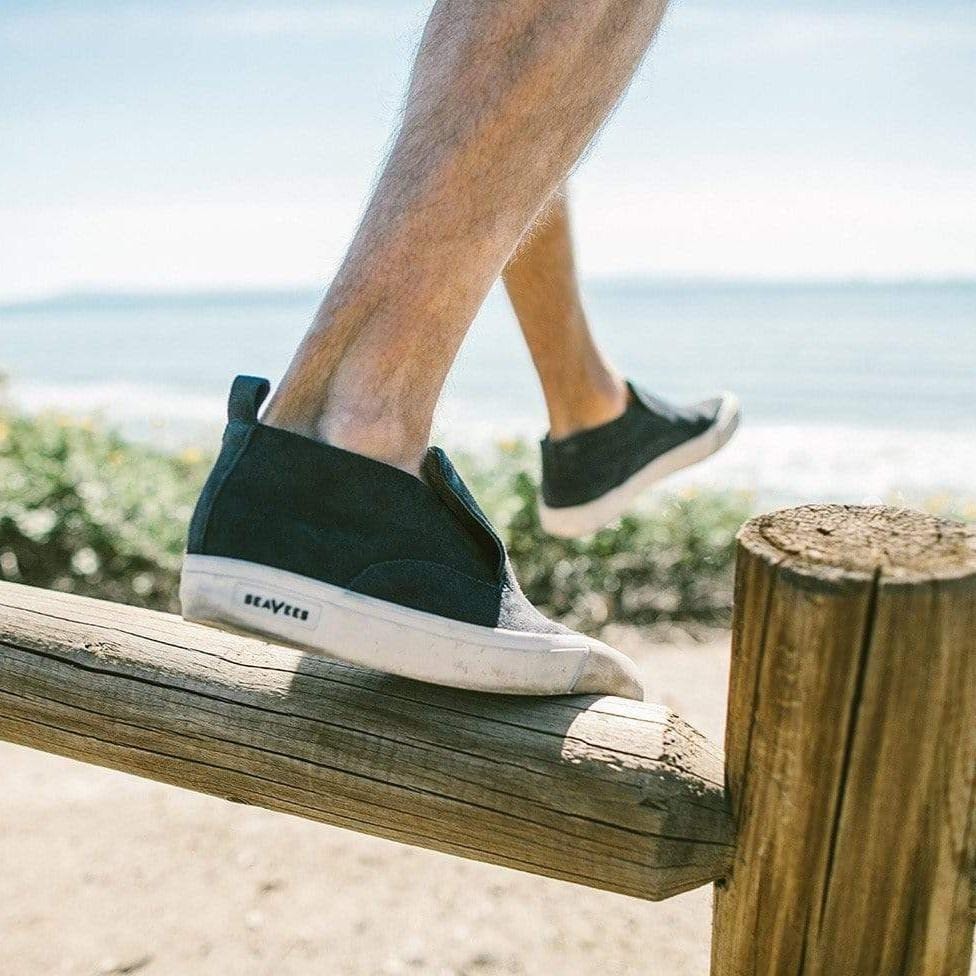 Men's Huntington Middie sneaker in Deep Navy suede, worn with shorts, one foot on a wooden beam by the ocean