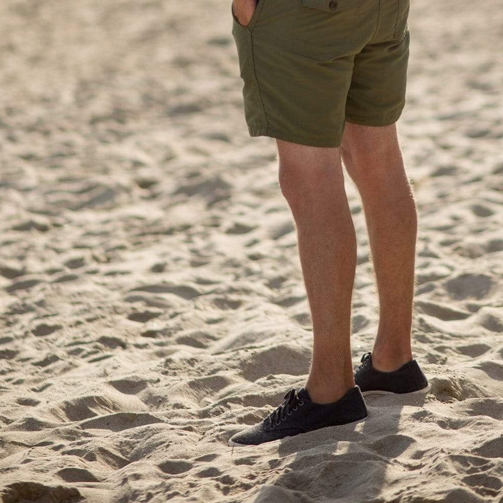 Men's Hermosa Sneaker in Black canvas, worn with olive shorts on sand