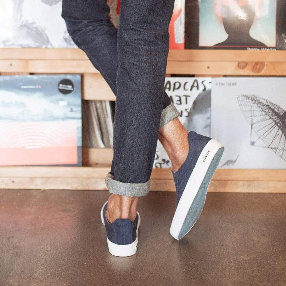 Men's Hawthorne Slip On in True Navy fabric, worn with cuffed dark jeans, shown from behind in front of record shelves