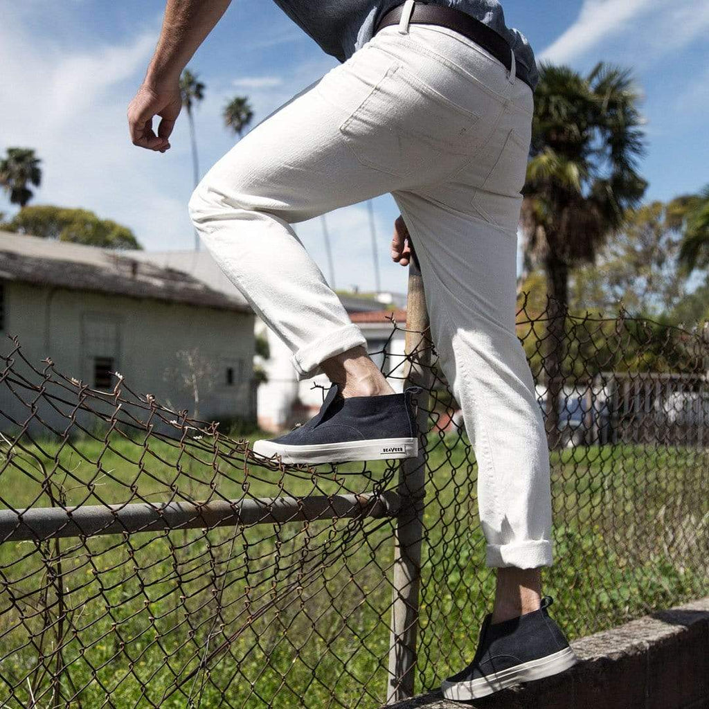 Men's Huntington Middie sneaker in Deep Navy suede, worn with white pants, stepping onto a chain-link fence