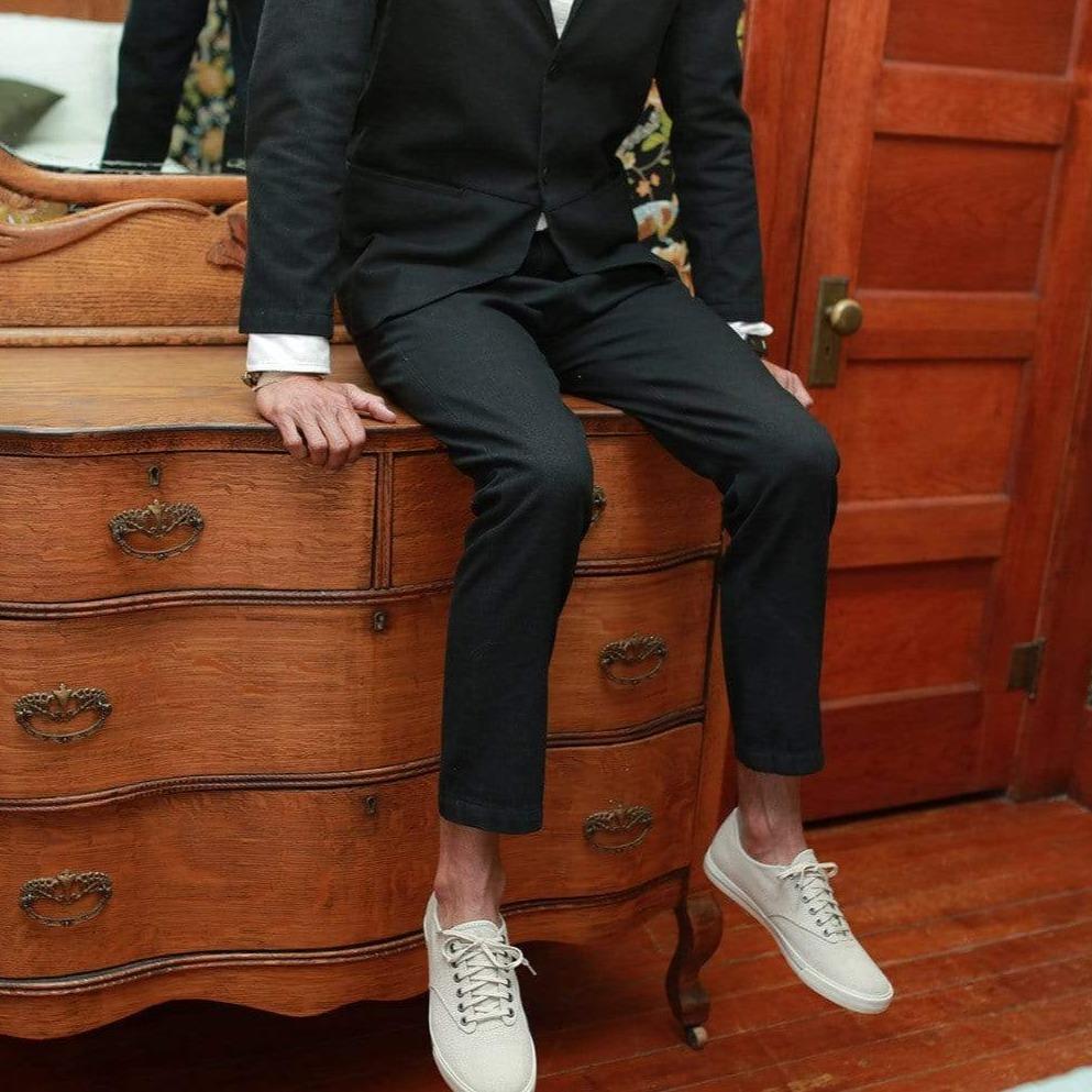 Men's Hermosa Sneaker in Natural canvas, worn with a dark suit by a person seated on a wooden dresser