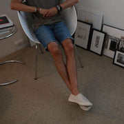 Men's Baja Slip On in Natural canvas, worn with denim shorts, seated in a studio chair