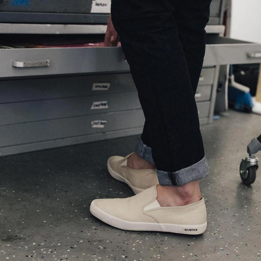 Men's Baja Slip On in Natural canvas, worn with cuffed black pants beside a flat file cabinet