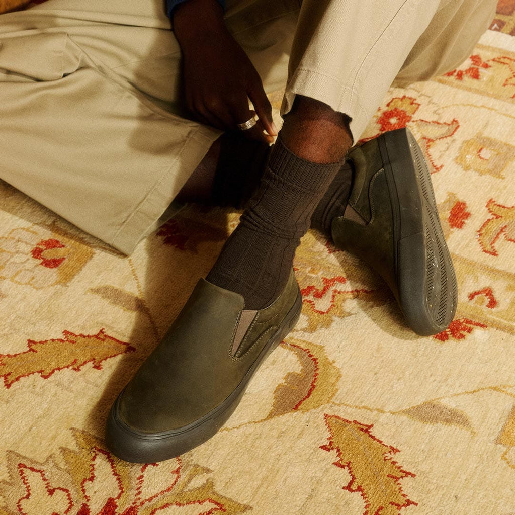 Men's Hawthorne Slip On in Black Olive leather, worn with khaki pants and dark socks on a patterned rug