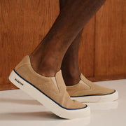 Men's Hawthorne Slip On in Camel suede, worn with bare ankles, one foot raised showing the navy stripe