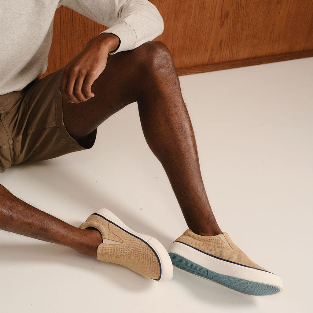 Men's Hawthorne Slip On in Camel suede, worn with khaki shorts, seated on a white floor