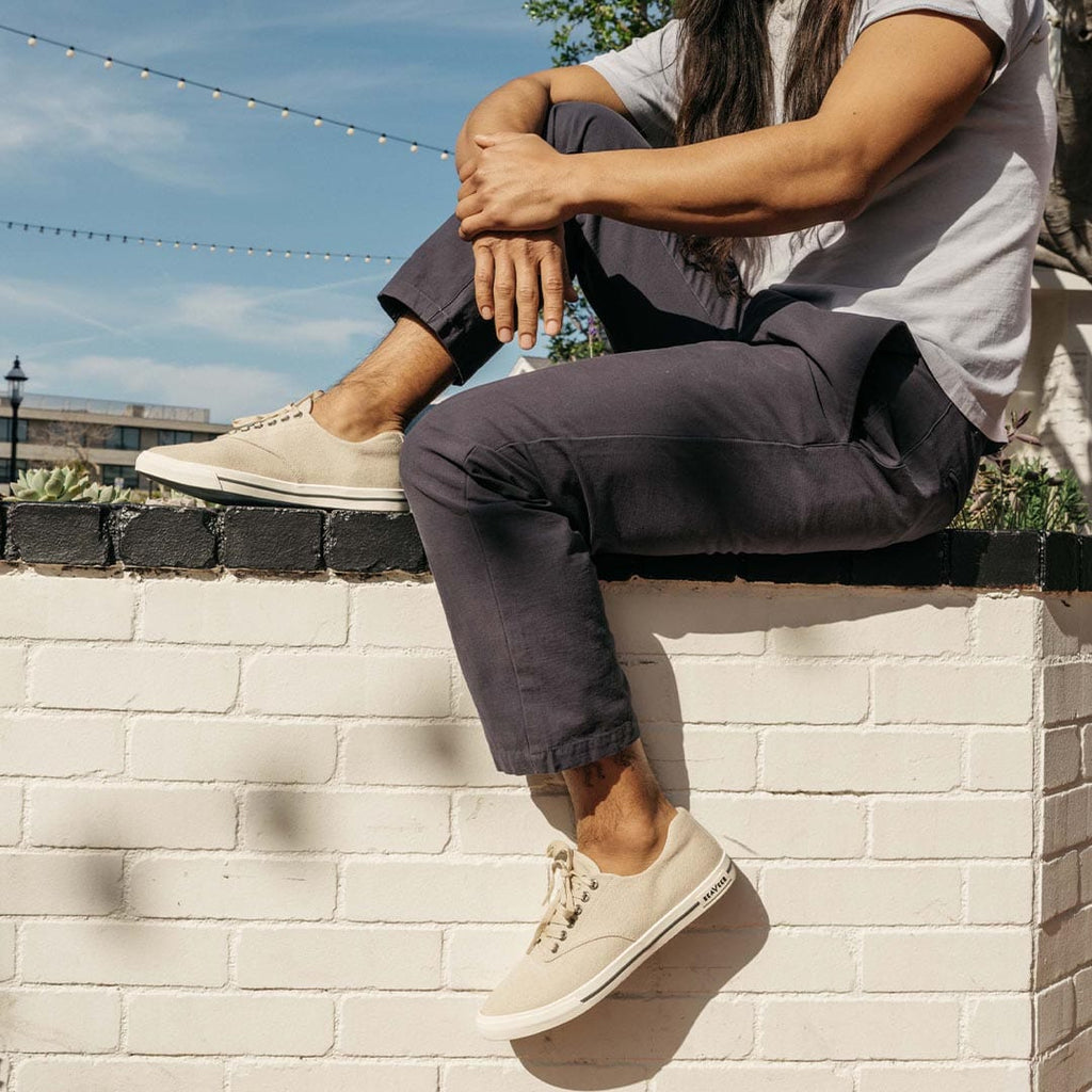 Men's Hermosa Sneaker in Natural canvas, worn with dark pants by a person seated on a white brick ledge