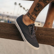 Men's Hermosa Sneaker in Slate Navy canvas, close-up with laces, foot resting on a wood rail by the beach
