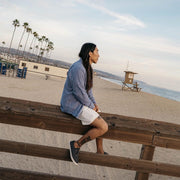Men's Hermosa Sneaker in Slate Navy canvas, worn by a person seated on a wooden beach rail