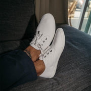 Men's Sixty Six Sneaker in White canvas, close-up with laces, worn with dark pants and feet propped on a couch