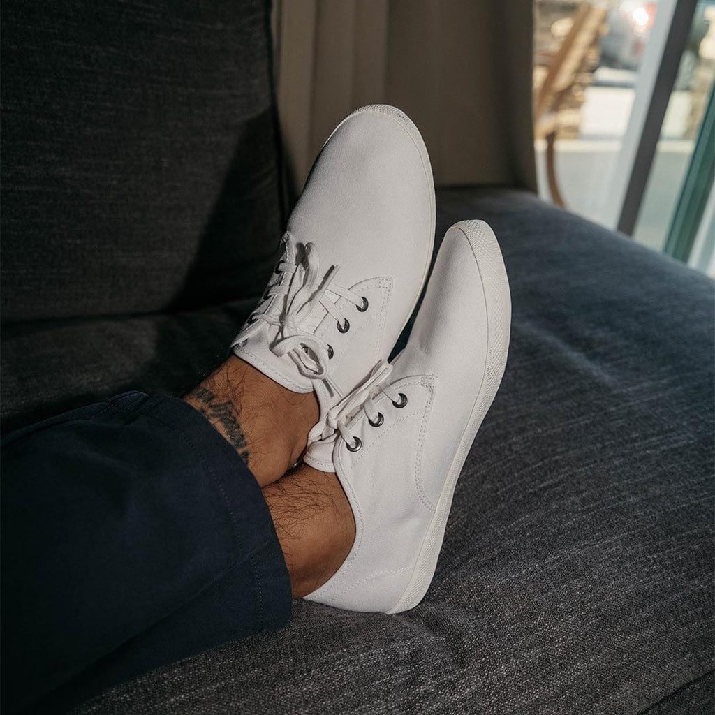 Men's Sixty Six Sneaker in White canvas, close-up with laces, worn with dark pants and feet propped on a couch