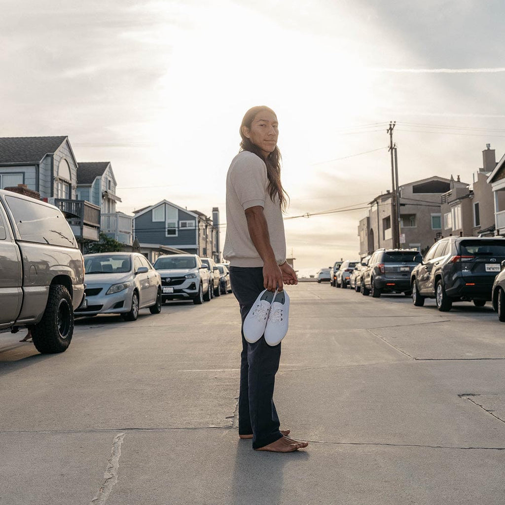 Men's Sixty Six Sneaker in White canvas, pair held in hand by a barefoot person standing in a street at sunset