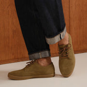 Men's Sun-tans Buck in Dusty Olive suede, worn with cuffed dark jeans on a white floor against wood paneling