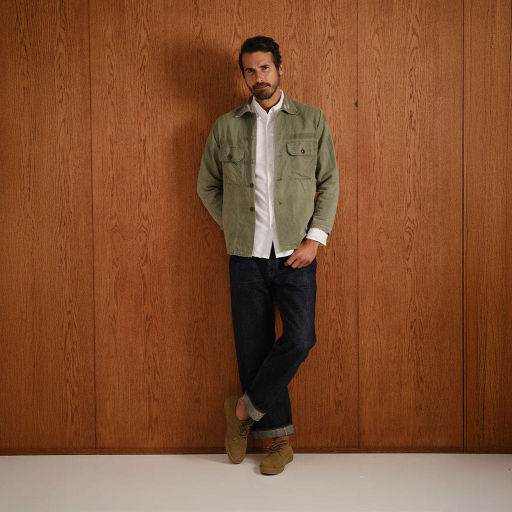 Men's Sun-tans Buck in Dusty Olive suede, worn by a person leaning on a wood-paneled wall in a green shirt