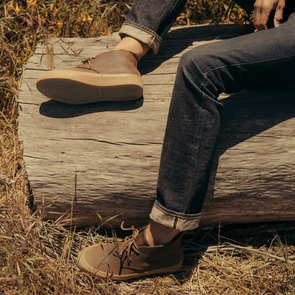 Men's Maslon Desert Boot in Kona leather, worn with dark jeans by a person seated on a log