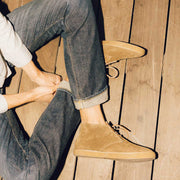 Men's Sun-tans Chukka in Desert Palm suede, worn with cuffed jeans by a person seated on a wood floor lacing the boot