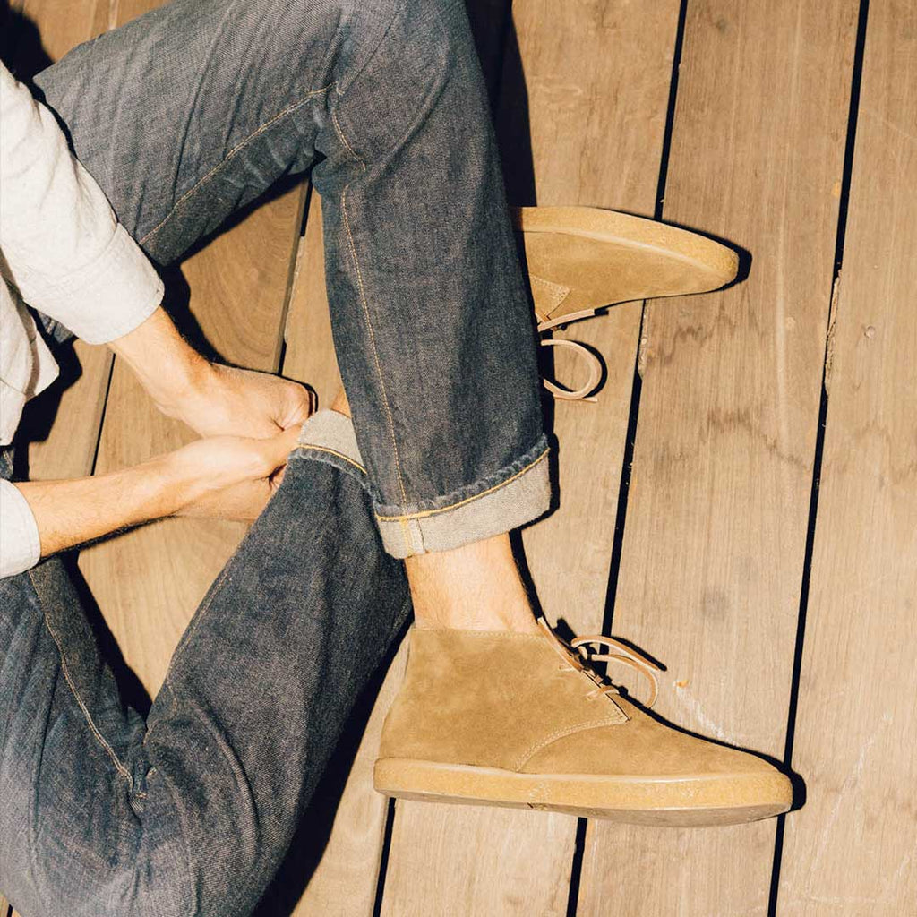 Men's Sun-tans Chukka in Desert Palm suede, worn with cuffed jeans by a person seated on a wood floor lacing the boot