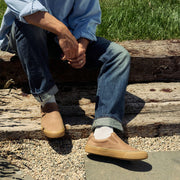 Men's Hawthorne Slip On in Sandstorm leather, worn with rolled jeans and white socks, seated on a wooden beam