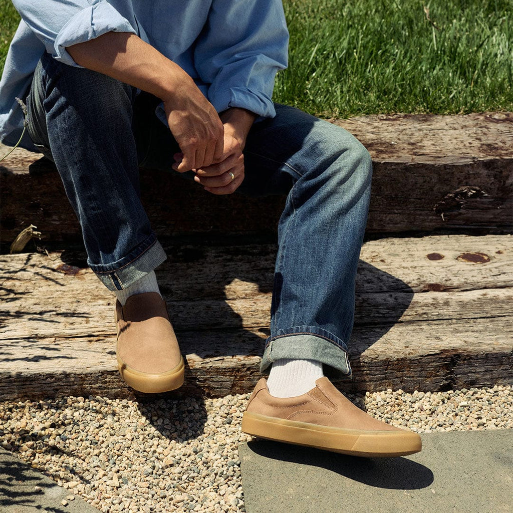 Men's Hawthorne Slip On in Sandstorm leather, worn with rolled jeans and white socks, seated on a wooden beam
