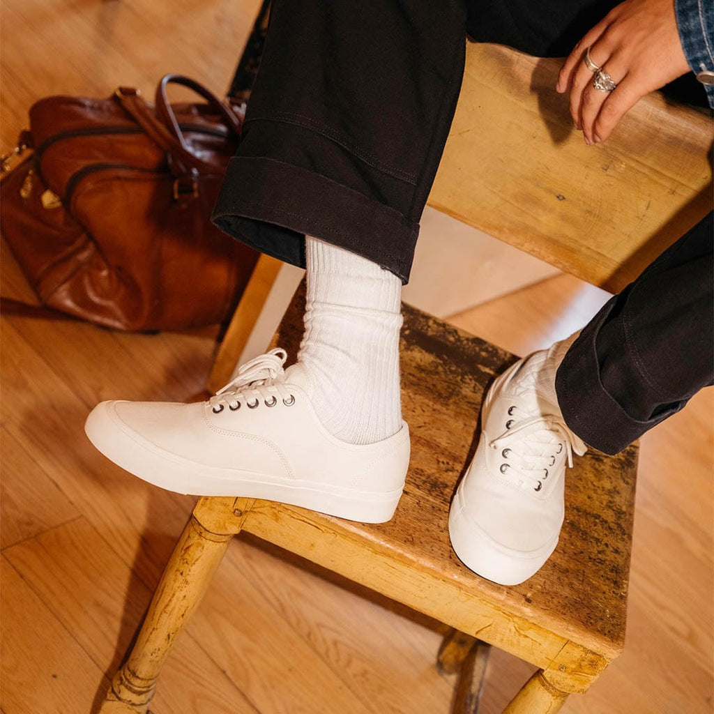 Men's Legend Sneaker in Bleach canvas, worn with white socks, feet resting on a wooden chair