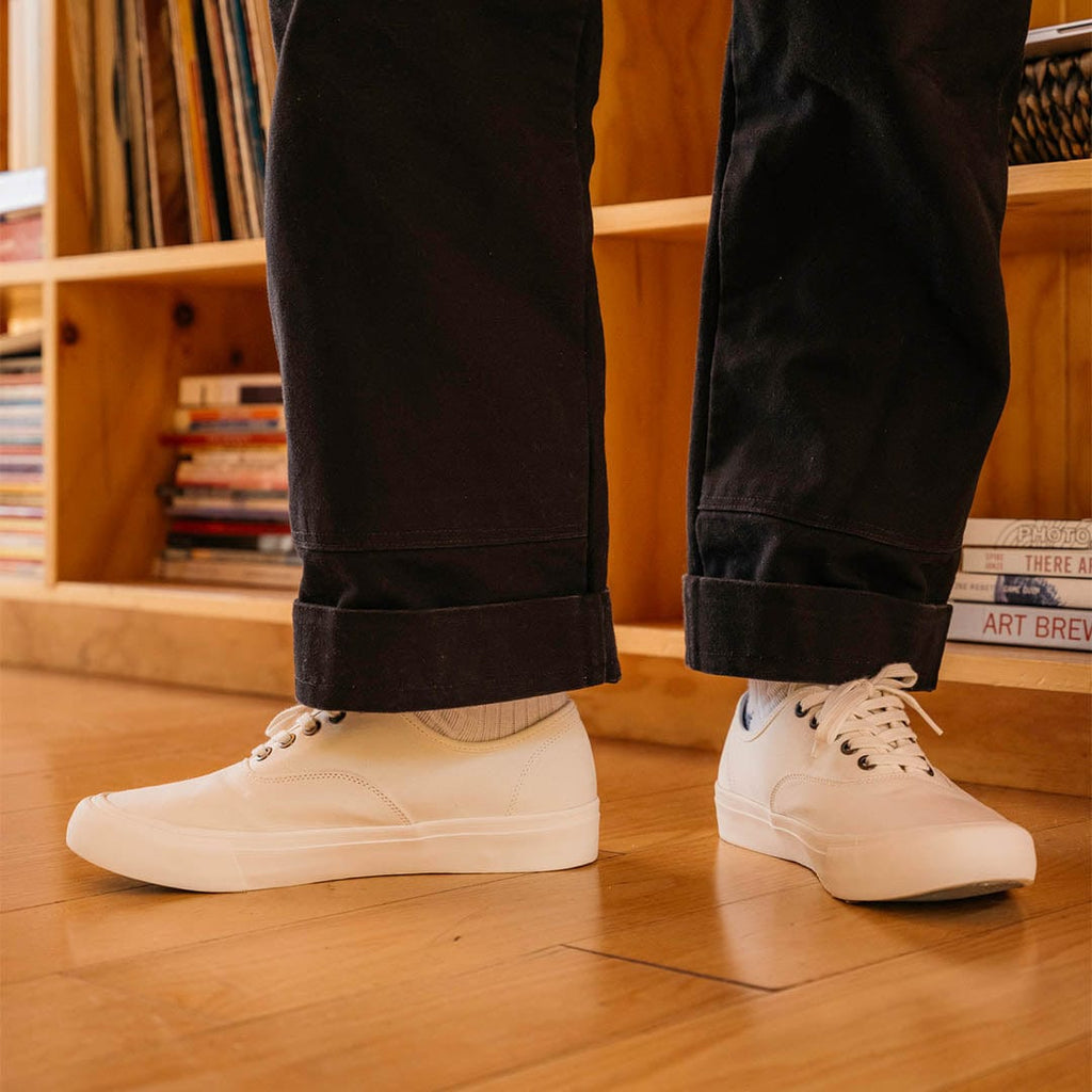 Men's Legend Sneaker in Bleach canvas, worn with dark cuffed pants in front of a wooden bookshelf