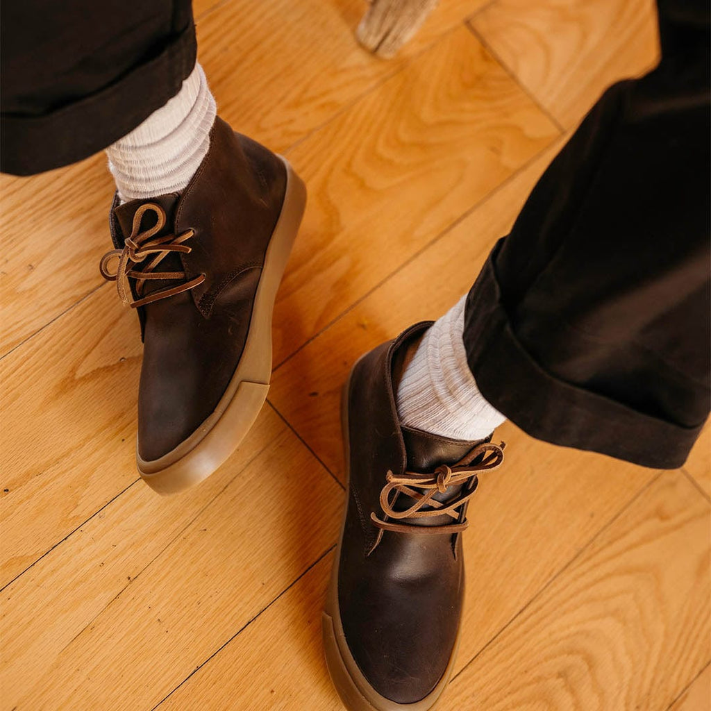 Men's Maslon Desert Boot in Kona leather, side close-up worn with white socks and dark pants on a wood floor