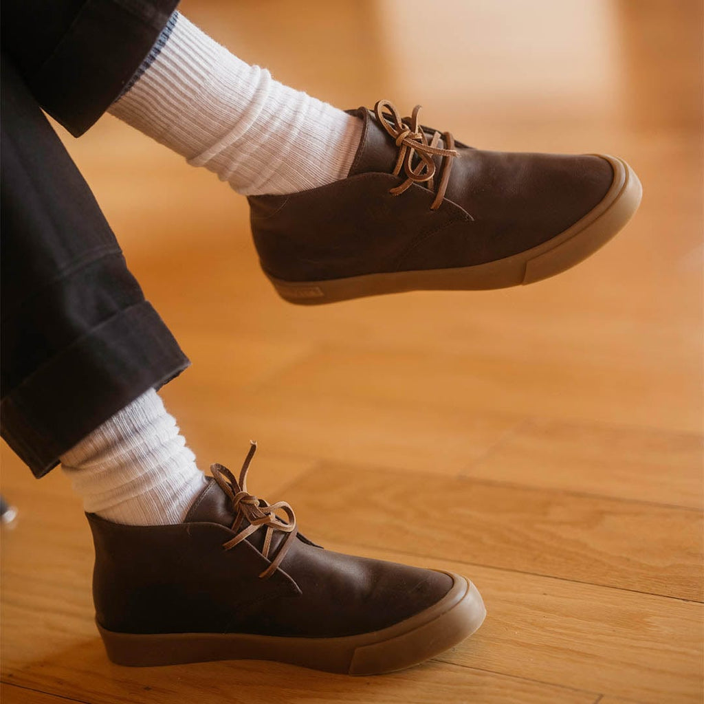 Men's Maslon Desert Boot in Kona leather, close-up worn with white socks, one foot crossed over the other, on a wood floor